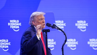President Trump is seen in profile as he gestures while speaking at a podium labeled "Annual Meeting Davos 2026." Behind him is a purple screen with "World Economic Forum" printed on it.