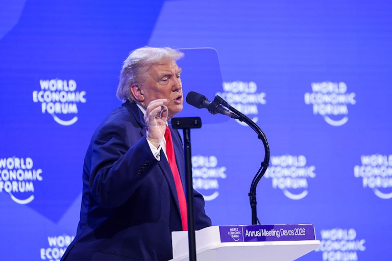President Trump is seen in profile as he gestures while speaking at a podium labeled "Annual Meeting Davos 2026." Behind him is a purple screen with "World Economic Forum" printed on it.