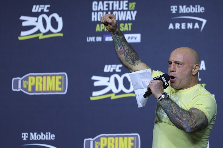 Joe Rogan holds up his fist while speaking into a hand-held microphone at a UFC event