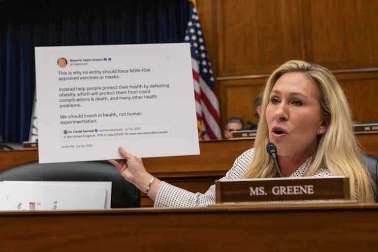 Marjorie Taylor Greene holds up a giant posterboard with one of her old tweets printed on it. It is about Covid, masks, vaccines, and obesity.