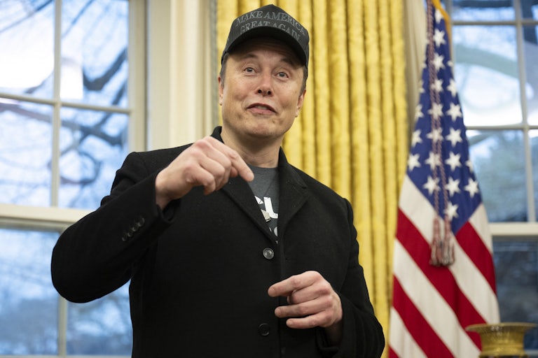 Elon Musk, wearing a black Make America Great Again hat and a black blazer over a t-shirt, stands in the Oval Office with an American flag and windows visible behind him.
