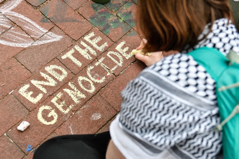A student protester wearing a keffiyeh writes "End the genocide" on the ground in chalk at the University of Michigan