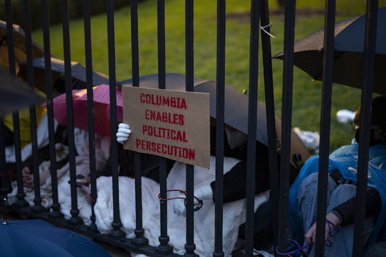 Someone holds a sign behind a gate reading "Columbia Enables Political Persecution." Several people, including the sign holder, crouch under umbrellas behind the gate.