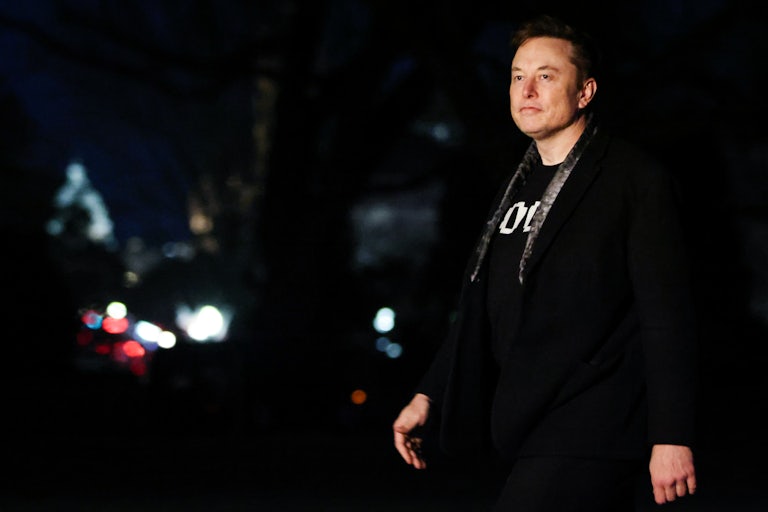Elon Musk walks outside the White House in the dark, while wearing a DOGE shirt.