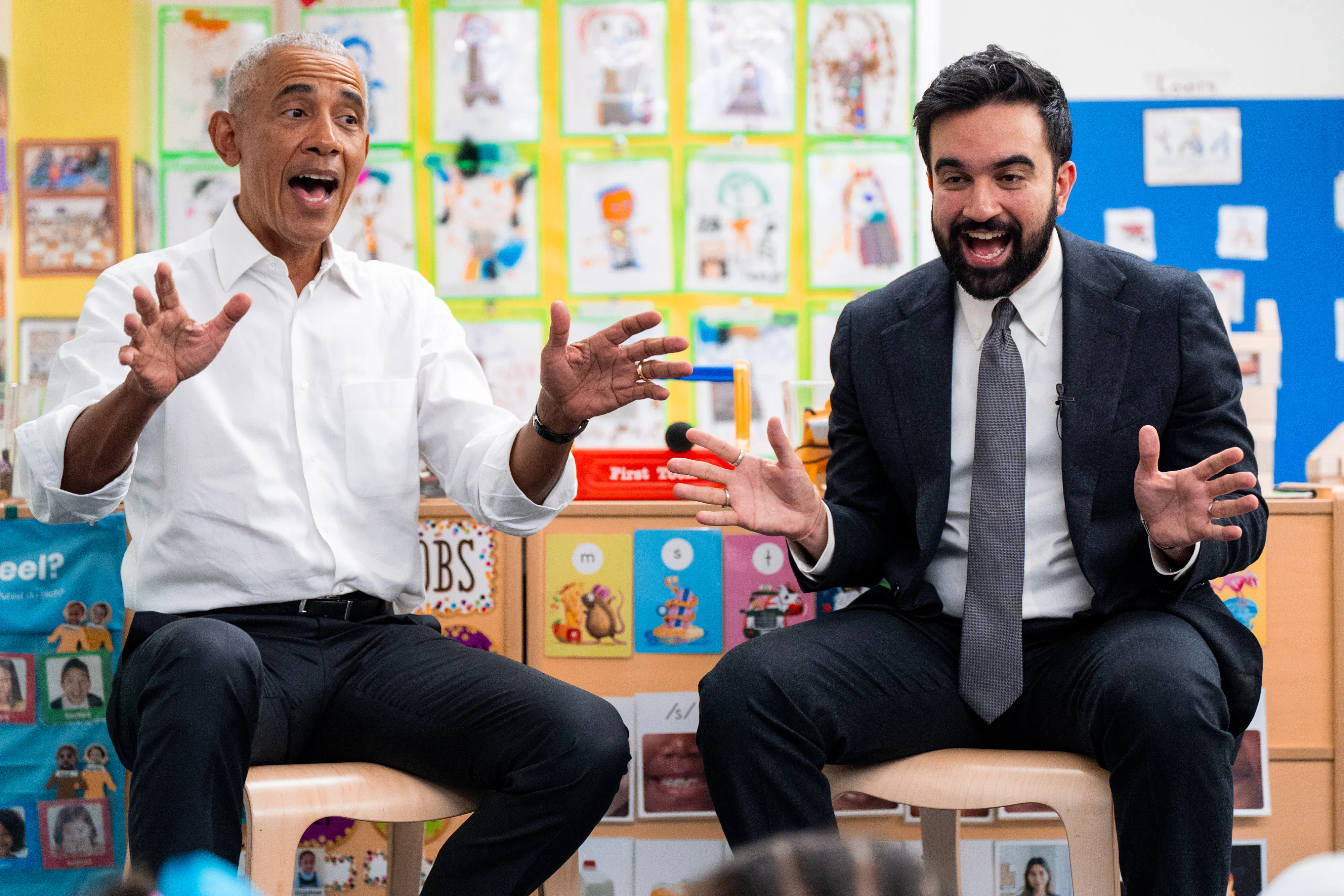 Barack Obama, wearing a white shirt, sits next to Zohran Mamdani in a preschool waving their hands with big, happy expressions.