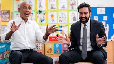 Barack Obama, wearing a white shirt, sits next to Zohran Mamdani in a preschool waving their hands with big, happy expressions.