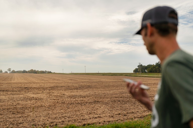 A farmer holds their phone while looking out at a soybean field