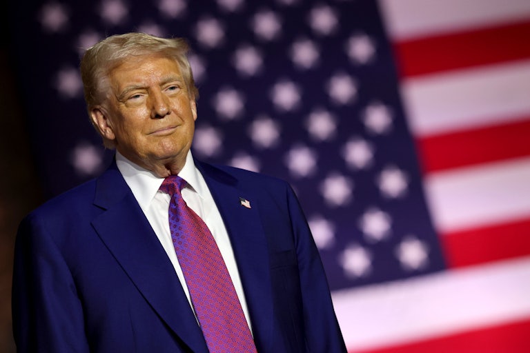 Donald Trump stands in front of a large U.S. flag and smiles slightly