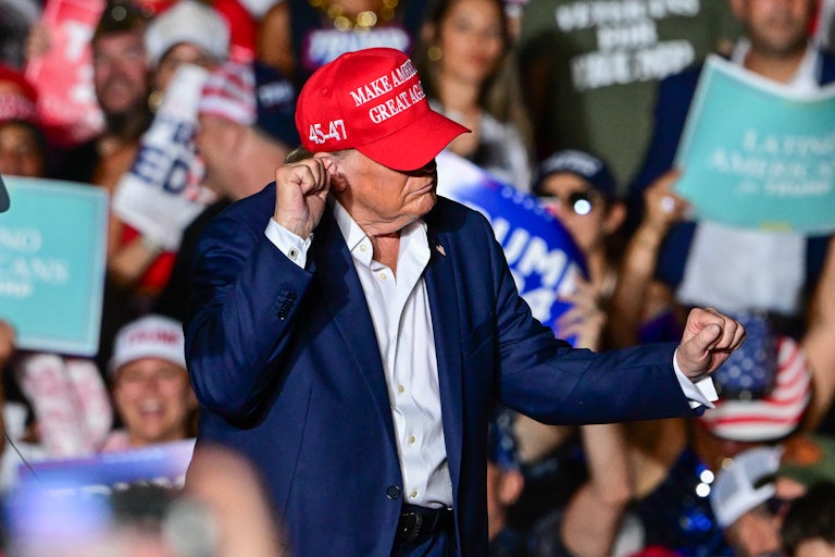 Donald Trump wears a MAGA cap and does a weird dance. A crowd of supporters can be seen in the background.