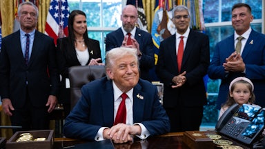 Donald Trump sits at his desk in the Oval Office of the White House, as RFK Jr. and others stand behind him.