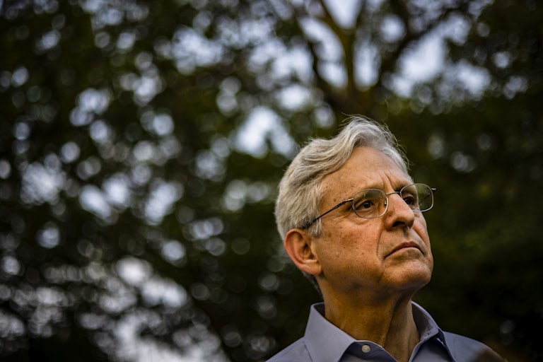 Attorney General Merrick Garland stands outside under some trees.