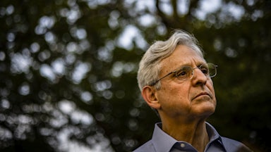 Attorney General Merrick Garland stands outside under some trees.