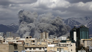 Smoke rises above buildings, with mountains in the background.