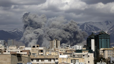 Smoke rises above buildings, with mountains in the background.