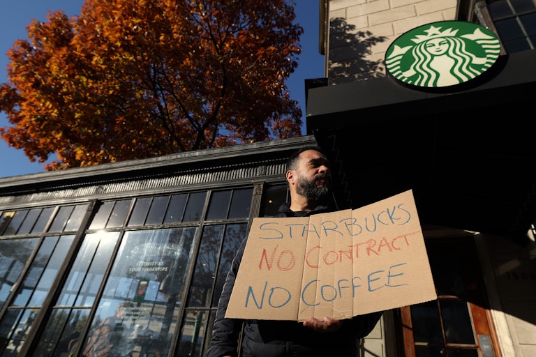 A man standing in front of a Starbucks holds a cardboard sign that reads "Starbucks No Contract No Coffee."