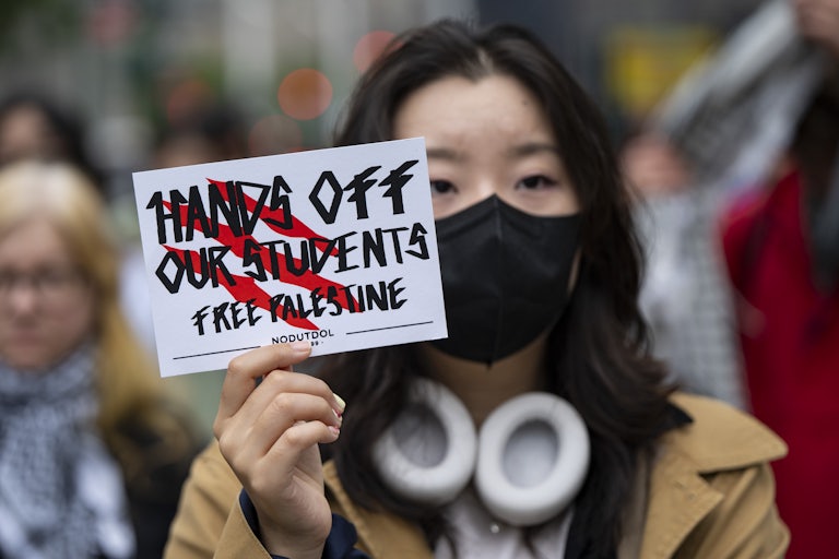 An Asian woman wearing a face mask holds a sign that reads "Hands Off Our Students Free Palestine."