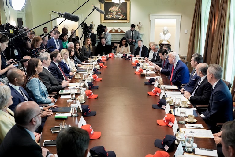 Trump and all his Cabinet members sit around a long conference table. Reporters stand with mics over their heads. On the table, each person has a red MAGA cap in front of them.