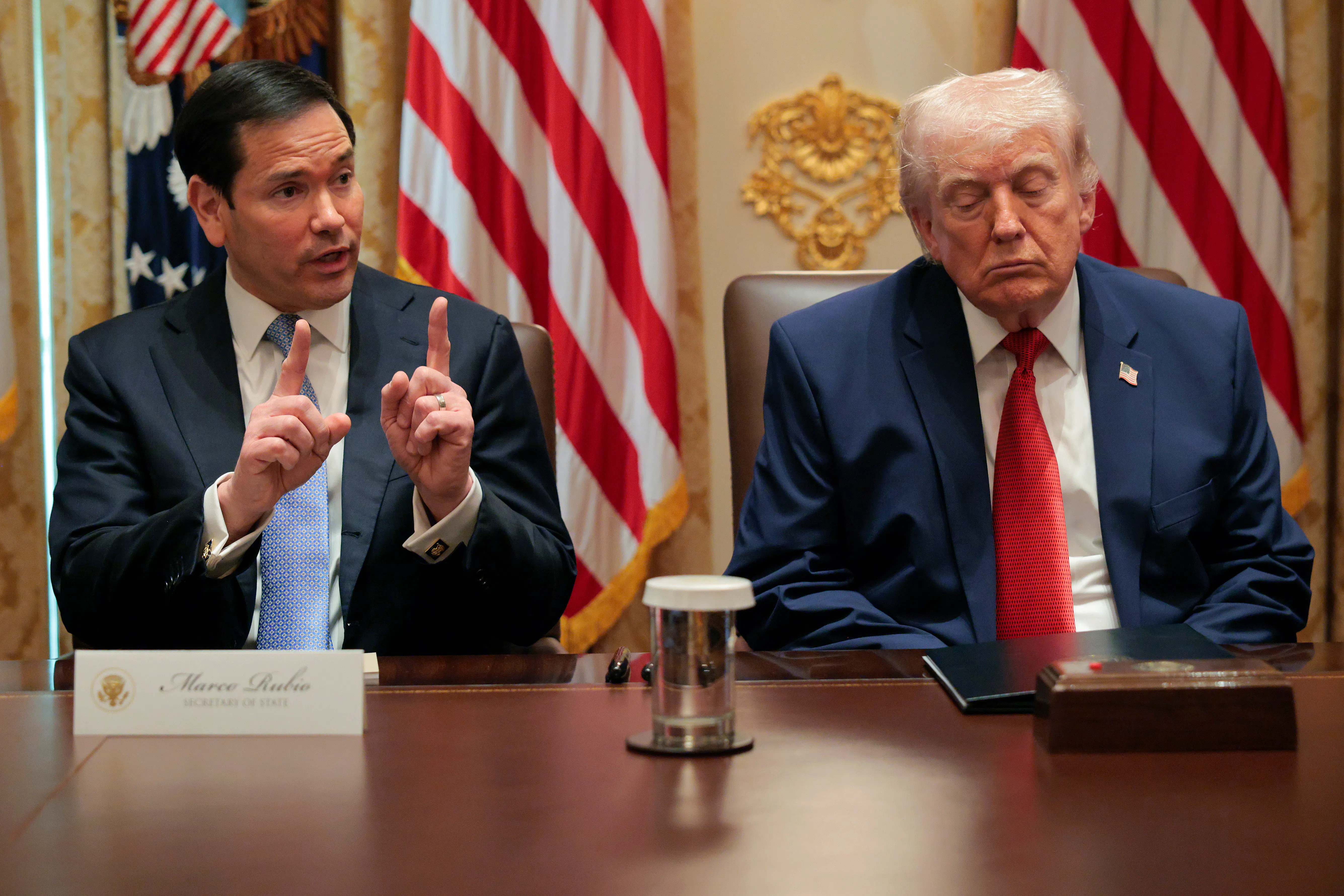 Marco Rubio, left, speaks while holding up both index fingers as Donald Trump, right, closes his eyes. Both are seated at a long wooden table. 