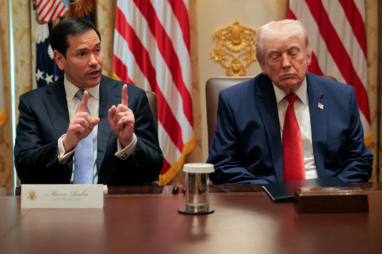 Marco Rubio, left, speaks while holding up both index fingers as Donald Trump, right, closes his eyes. Both are seated at a long wooden table.