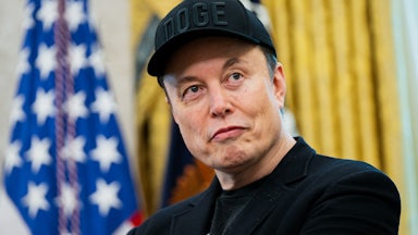 Elon Musk wears a black DOGE cap and smiles weirdly while sporting a black eye in the Oval Office of the White House.