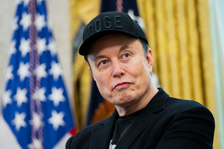 Elon Musk wears a black DOGE cap and smiles weirdly while sporting a black eye in the Oval Office of the White House.