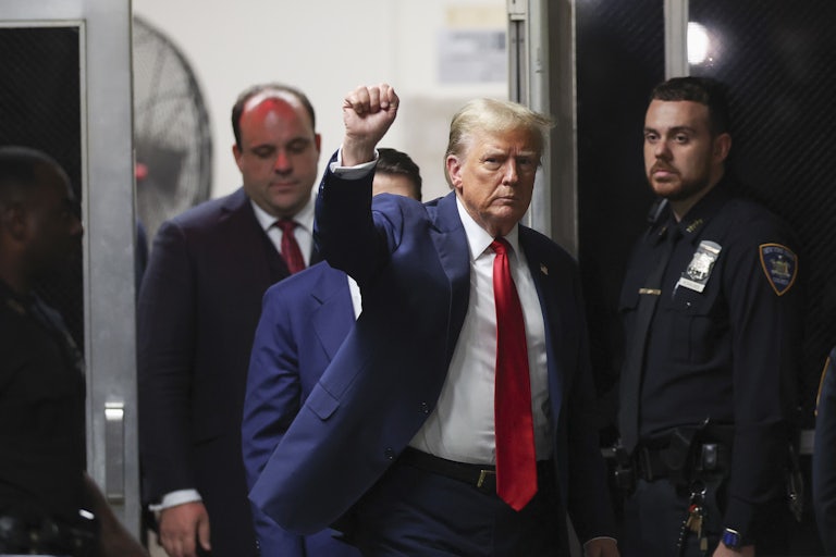 Donald Trump, walking in court, looks to the camera and raises his right fist, as if in victory. Boris Epshteyn is behind him. A security guard stands next to Trump.