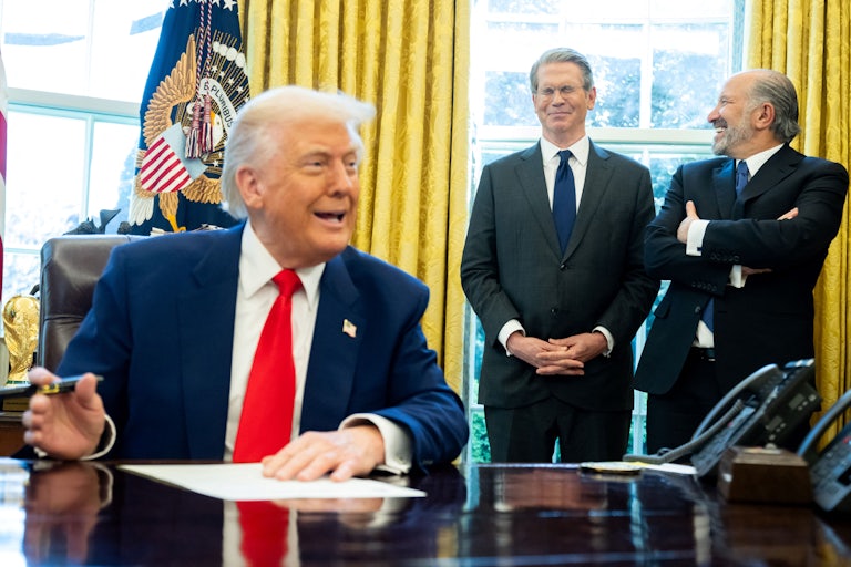 Donald Trump speaks to reporters while sitting at his desk in the Oval Office. Treasury Secretary Scott Bessent and Commerce Secretary Howard Lutnick stand behind him and laugh.