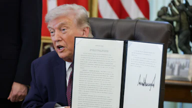 President Donald Trump holds up a signed executive order while sitting at his desk in the Oval Office of the White House.