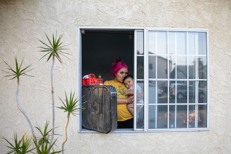 A woman with pink hair, seen from the outside of her building, stands at the window of her apartment in front of a fan holding a baby. A palm tree is seen next to the window.