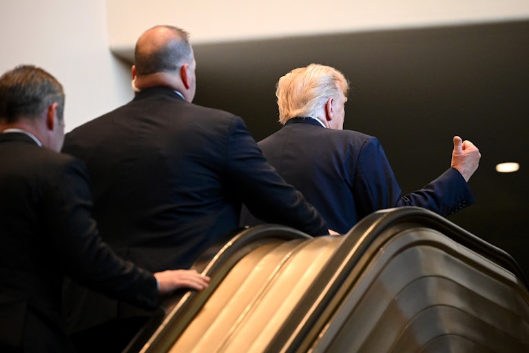 Donald Trump gives a thumps up (or is pointing behind him) as he gets off the escalator at the United Nations. Two men walk up the escalator behind him.