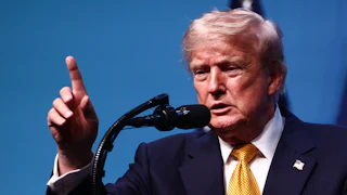 Donald Trump raises a finger while speaking into a microphone.