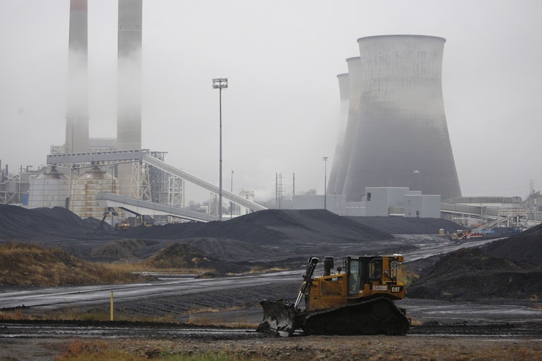 A bulldozer drives in front of coal piles and cooling towers.
