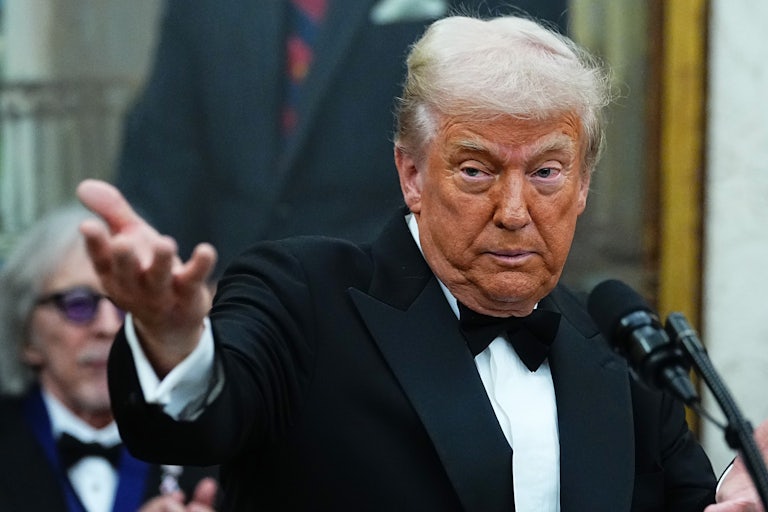 Donald Trump wears a tux and raises a hand outward while speaking at a mic.