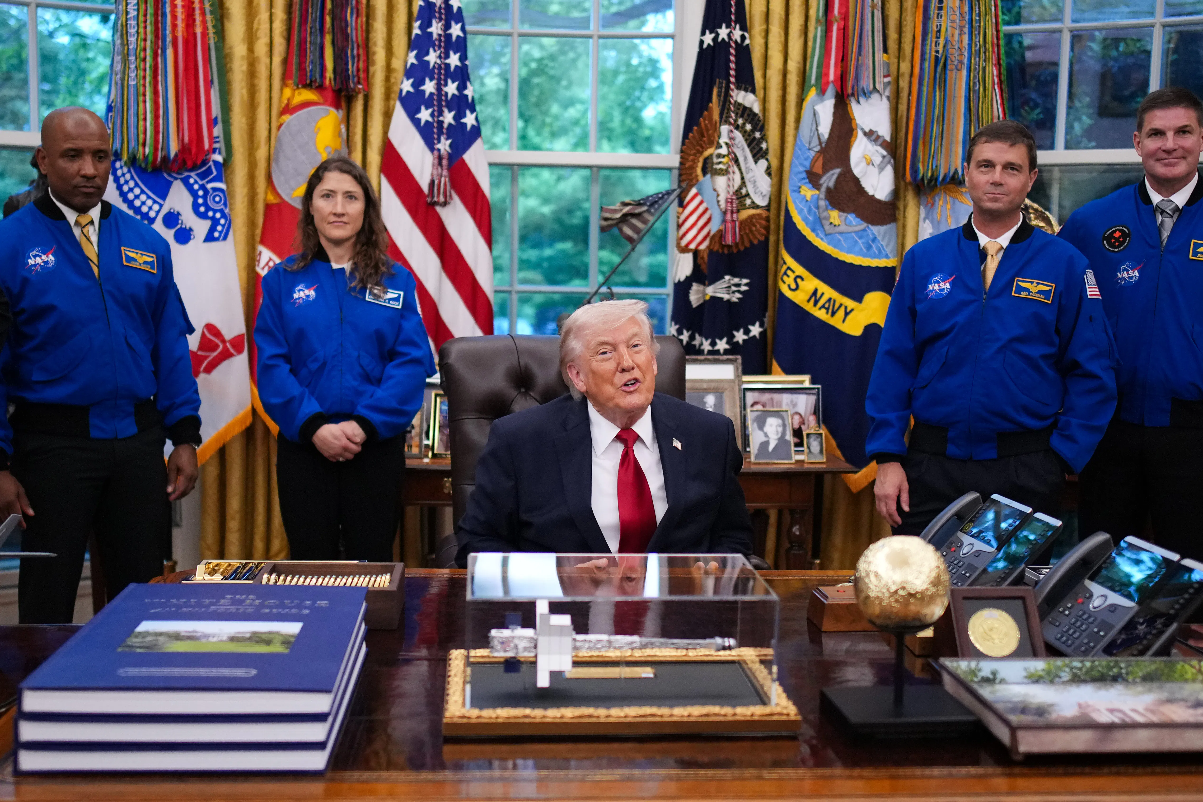 Donald Trump speaks while sitting at his desk in the Oval Office. He is flanked by the astronauts from the Artemis II mission.
