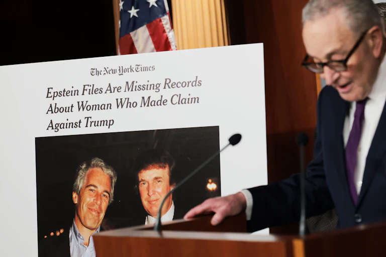 Senator Chuck Schumer speaks at a podium next to a poster-sized printout of a New York Times article. The article has a photo of Donald Trump and Jeffrey Epstein and is headlined, "Epstein Files Are Missing Records About Woman Who Made Claim Against Trump"
