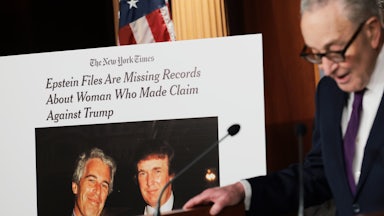 Senator Chuck Schumer speaks at a podium next to a poster-sized printout of a New York Times article. The article has a photo of Donald Trump and Jeffrey Epstein and is headlined, "Epstein Files Are Missing Records About Woman Who Made Claim Against Trump"