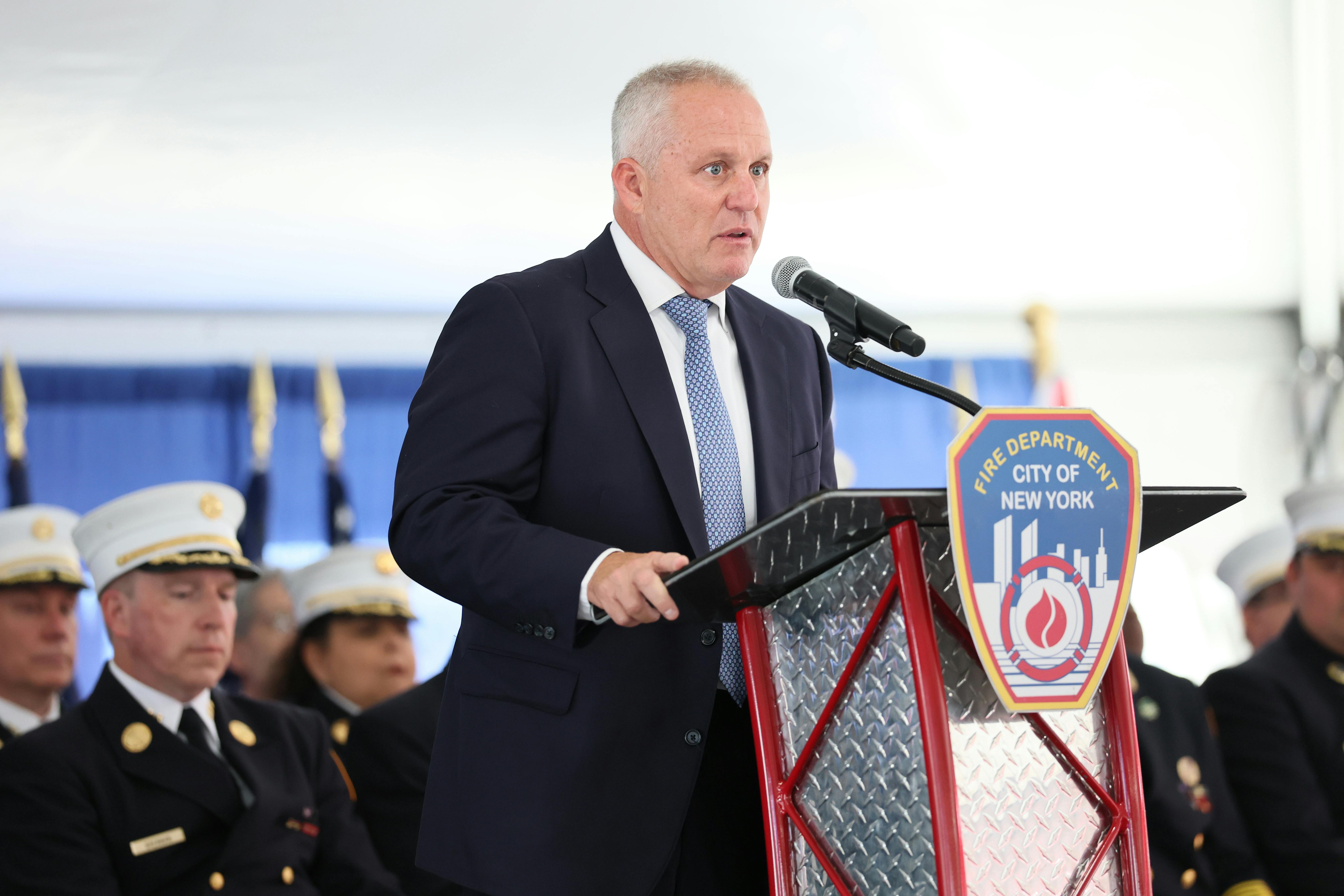 FDNY Fire Commissioner Robert Tucker speaks on stage behind a lectern that reads "Fire Department City of New York," while others speak behind him.