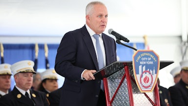 FDNY Fire Commissioner Robert Tucker speaks on stage behind a lectern that reads "Fire Department City of New York," while others speak behind him.