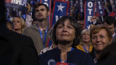 A women stands with her hands clasped in front of a crowd of onlookers watching former First Lady Michelle Obama deliver a speech on night two of the 2024 Democratic National Convention.