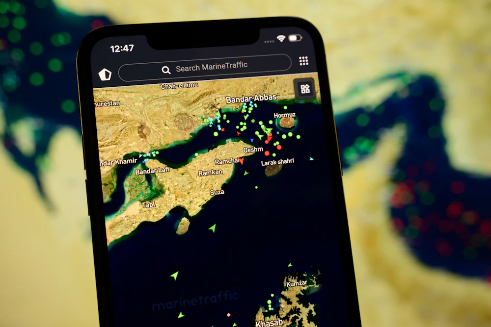 A smartphone displays the MarineTraffic app showing numerous ship beacons near the Strait of Hormuz with a satellite view in the background on April 8, 2026.