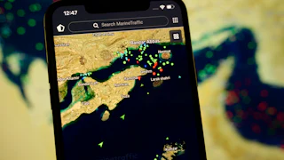 A smartphone displays the MarineTraffic app showing numerous ship beacons near the Strait of Hormuz with a satellite view in the background on April 8, 2026.