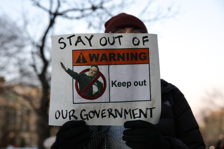 A protester holds a sign blocking his face that reads "Stay Out of Our Government, Warning Keep Out" With a picture of Elon Musk doing the Hitler salute