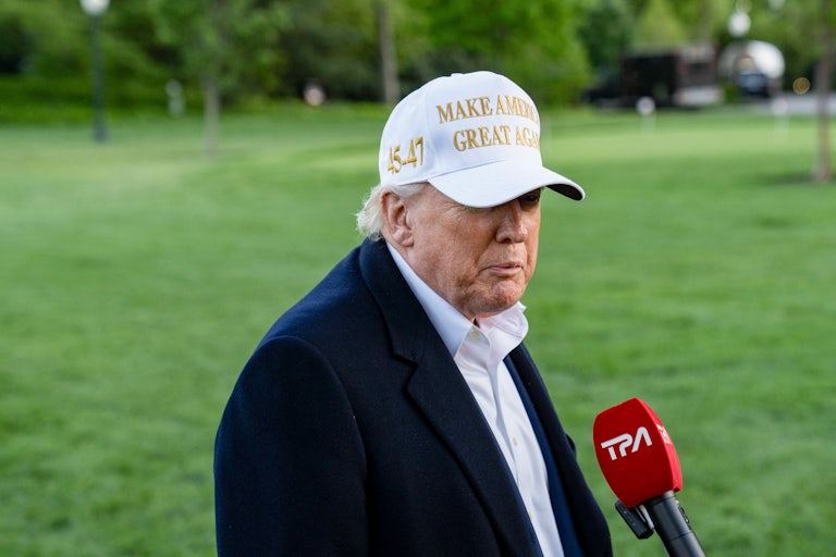 Donald Trump purses his lips while speaking into a microphone. He wears a white "Make America Great Again" hat