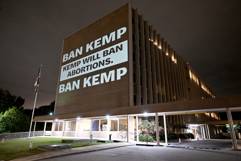 A projection on a building reads "BAN KEMP" and "KEMP WILL BAN ABORTIONS."