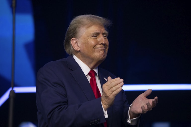 Donald Trump smiles weirdly and claps. You can see the outline of his spray tan on his face.