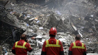 Three men in uniform stand in front of a collapsed building.