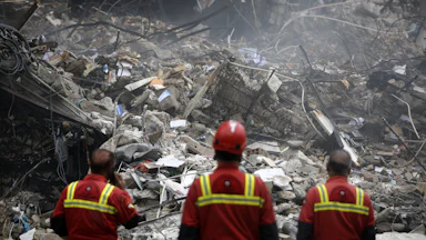 Three men in uniform stand in front of a collapsed building.