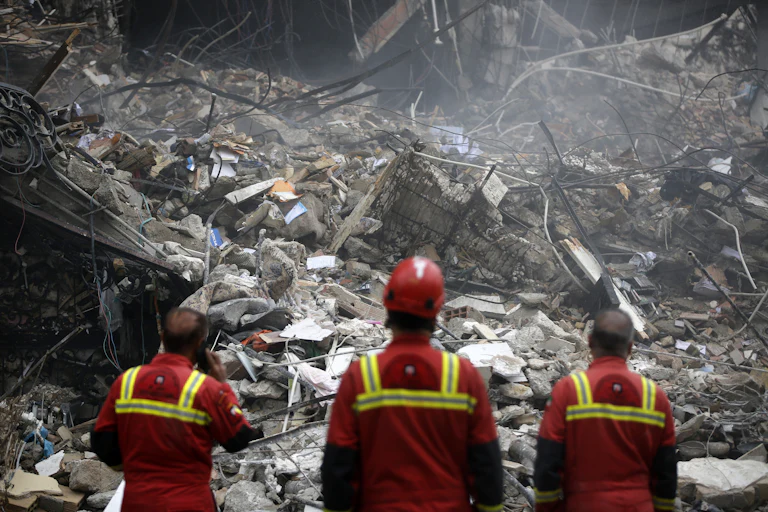 Three men in uniform stand in front of a collapsed building.