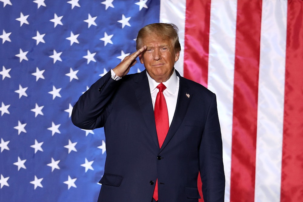 Donald Trump salutes while standing in front of a massive American flag