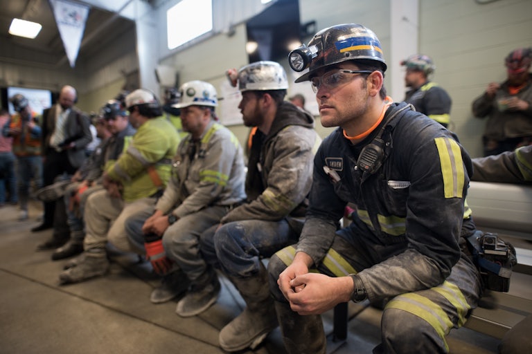Coal miners sit in a room on chairs waiting.